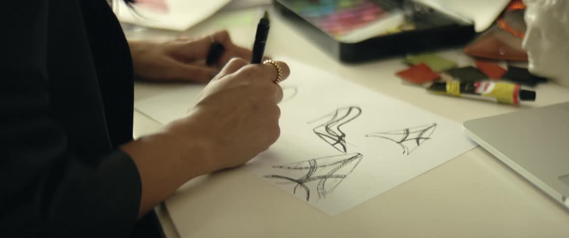 A woman in a room by a table drawing on a piece of paper next to a laptop with a wall in the background