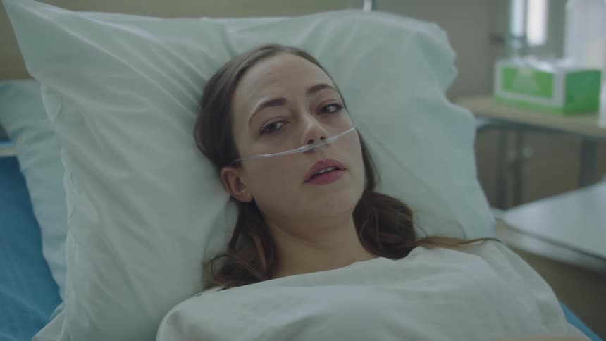 A woman is laying in a hospital bed on oxygen while a nurse moves a cup in the background
