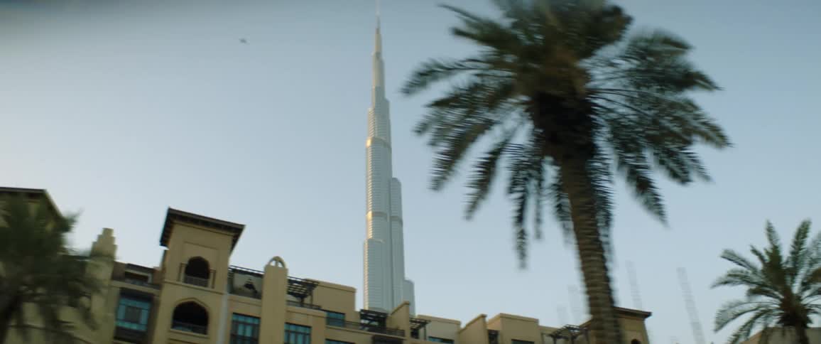 A building with palm trees in the foreground with a skyscraper the sky and clouds in the background