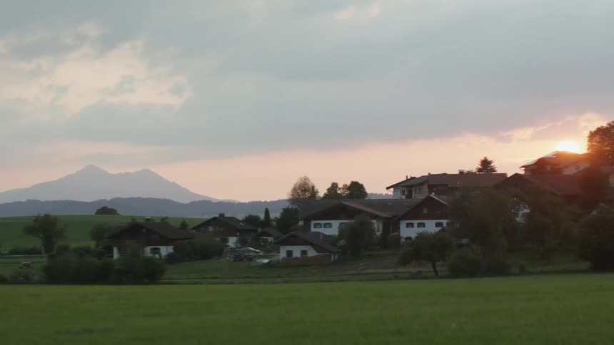 Houses and trees next to fields with cloudy mountains in the background at sunset