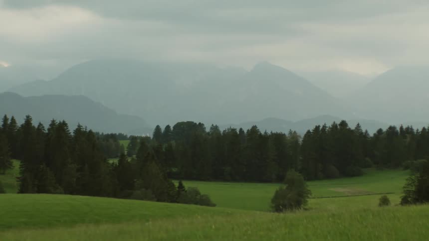 Clouds cover rolling green hills and a forest area with mountains in the background
