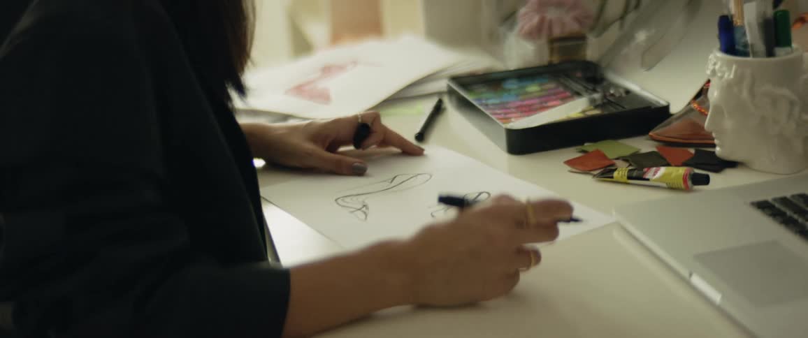 Woman sits at desk near laptop drawing on white paper