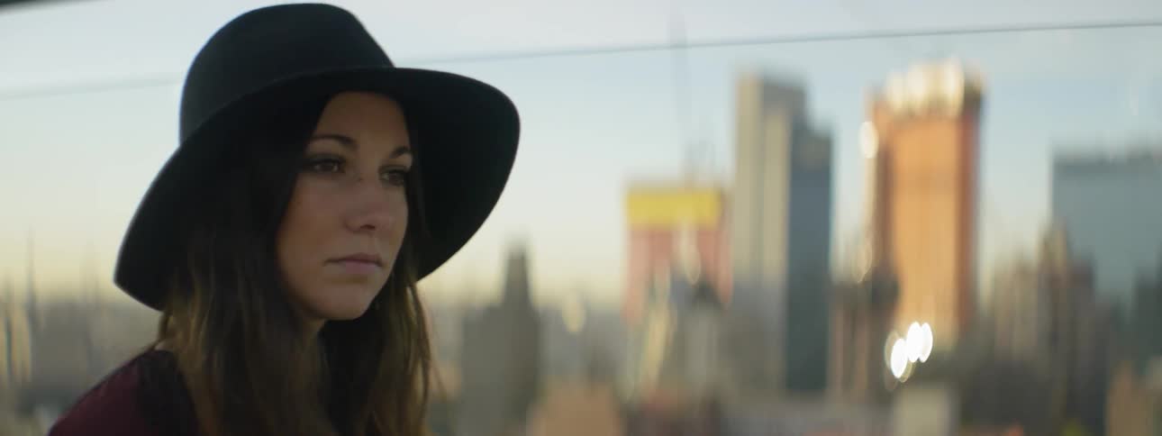 Woman in hat staring off with large city buildings behind her