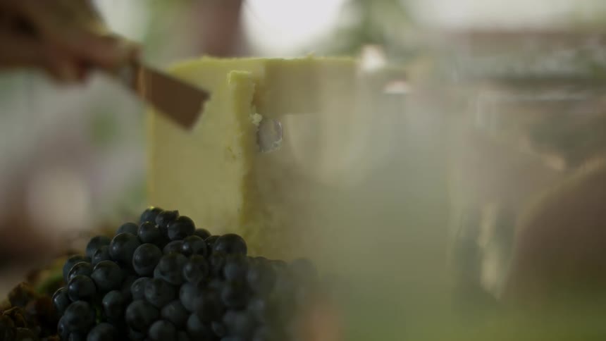 A person in a room cutting cheese with a knife with grapes in the foreground