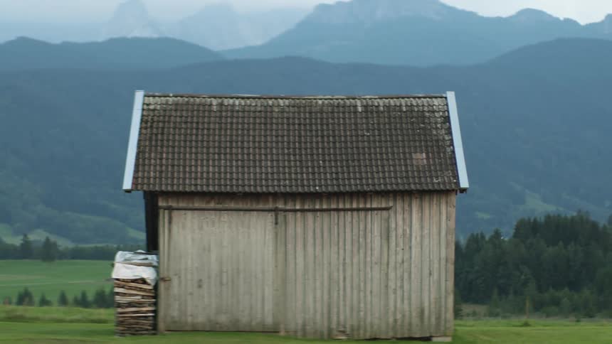 An old wooden building sits on land with trees nearby and mountains in the distance