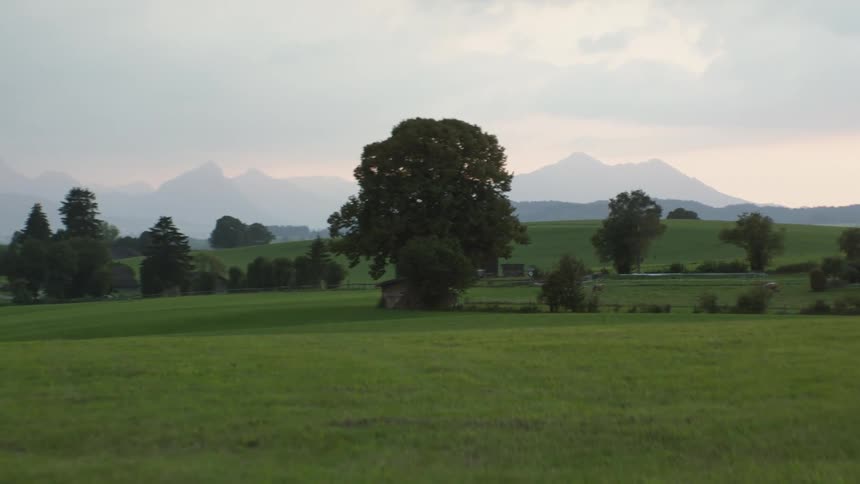 Fields stretch past buildings to hills and misty mountains on foggy horizon under red tinted sky