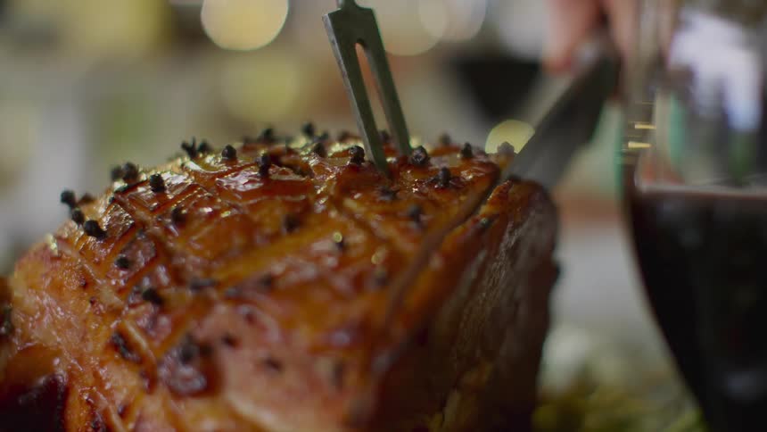 Person slices into ham using knife and two tine fork as near by wine in wine glass sloshes