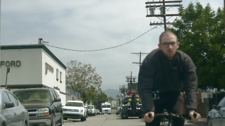 A man wearing glasses is riding a bicycle on a street and moving past parked cars and buildings