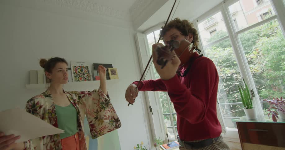 A man and a woman in a room standing still with the man holding a violin with a wall and window in the background