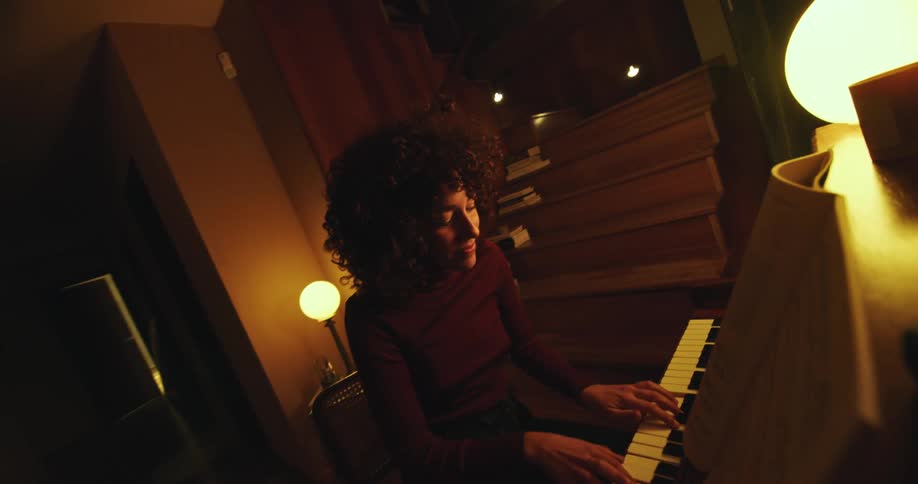 Young woman with curly hair playing piano inside a dimly lit room at home