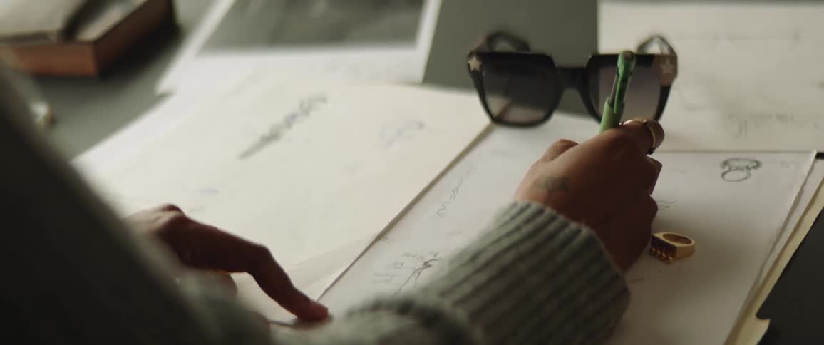 Woman sits sketching jewelry designs on paper as gold ring sits nearby