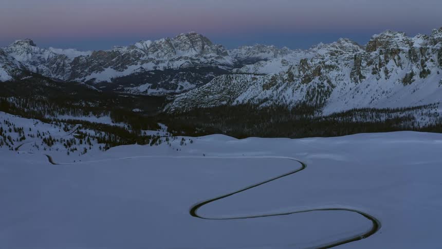 Aerial view of winding road in snowy field with mountain ranges and forest