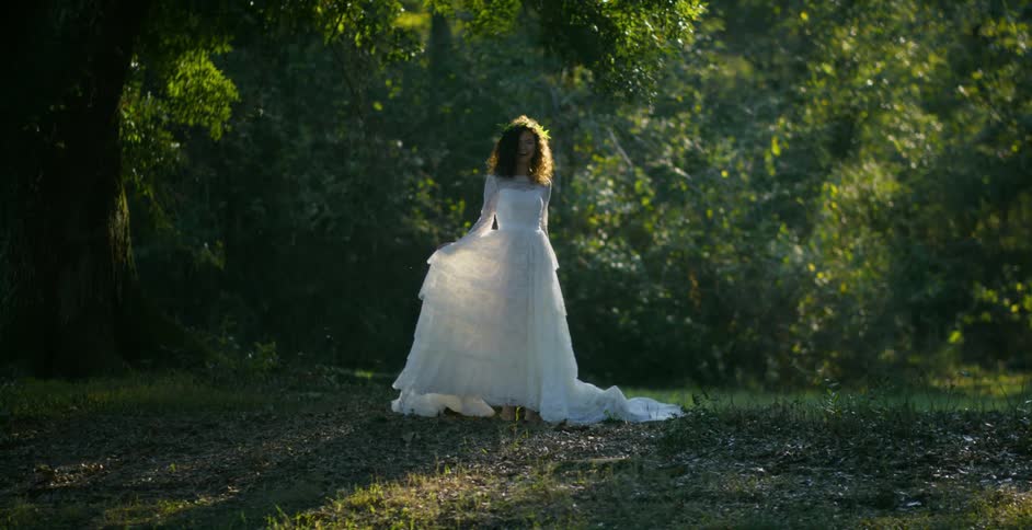 Woman with curly hair and wedding dress stands next to tree posing for the camera in the forest
