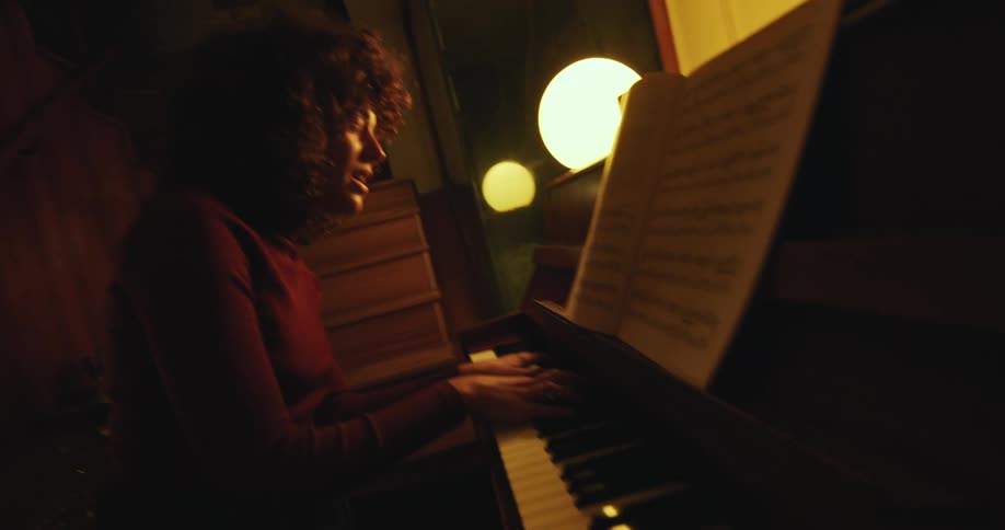 Woman with curly hair and red sweater plays piano and sings song in dim lit room