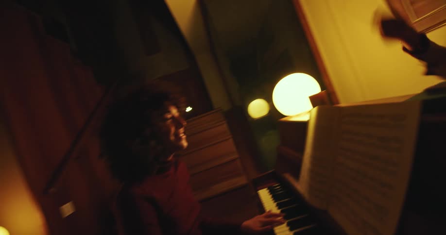 In a dimly lit room a mother happily plays an upright piano while her son stands behind it energetically conducting with writing instruments