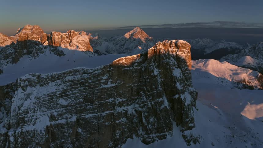 Snow covers land around mountains including cliff as sun casts shadows across area