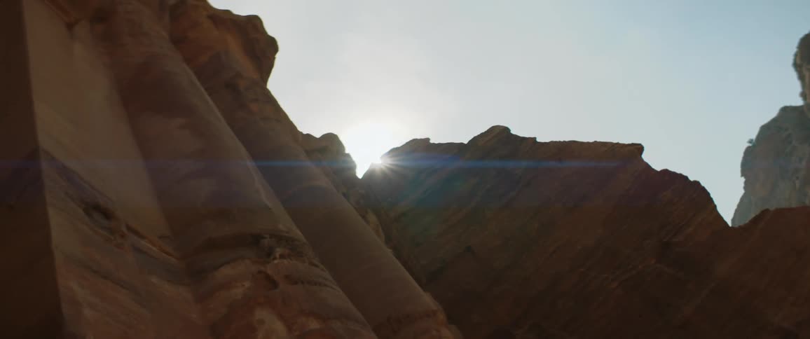Sun shines from top of red cliff near columns carved partly out of rock face