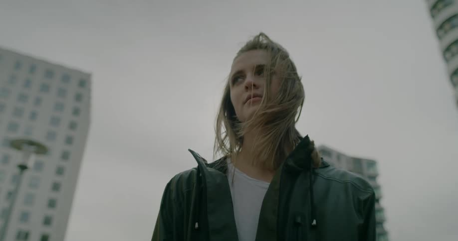 A woman wearing a dark colored jacket is standing in town with tall buildings behind her as the wind blows her hair