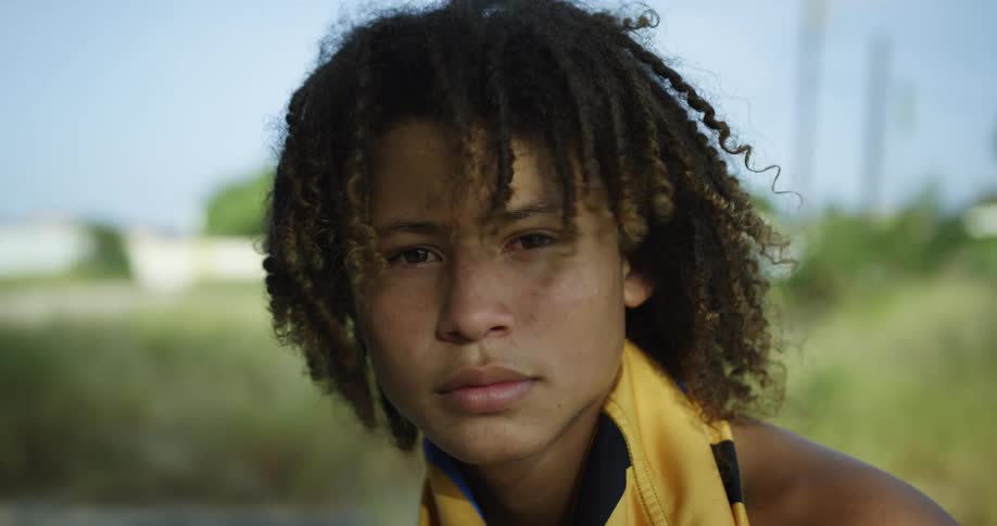 Shirtless boy with long curly hair and yellow shirt around neck looks at camera on windy day
