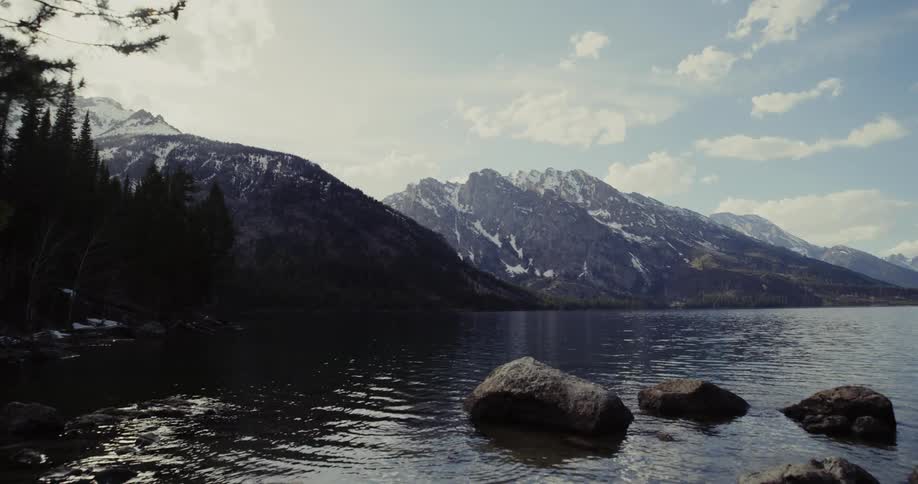 Water forms ripples around boulders near shore as it reflects conifer trees bordering snow covered mountains under a cloudy sky