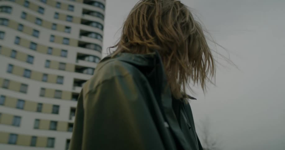Woman wearing light hooded jacket stands on windy day near apartment buildings