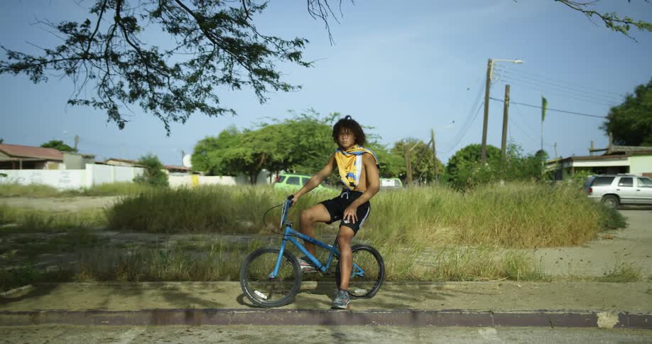 Youth sits on bicycle on sidewalk near overgrown lot