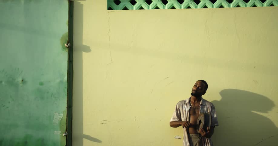 Man resting against a wall playing a makeshift musical instrument at evening