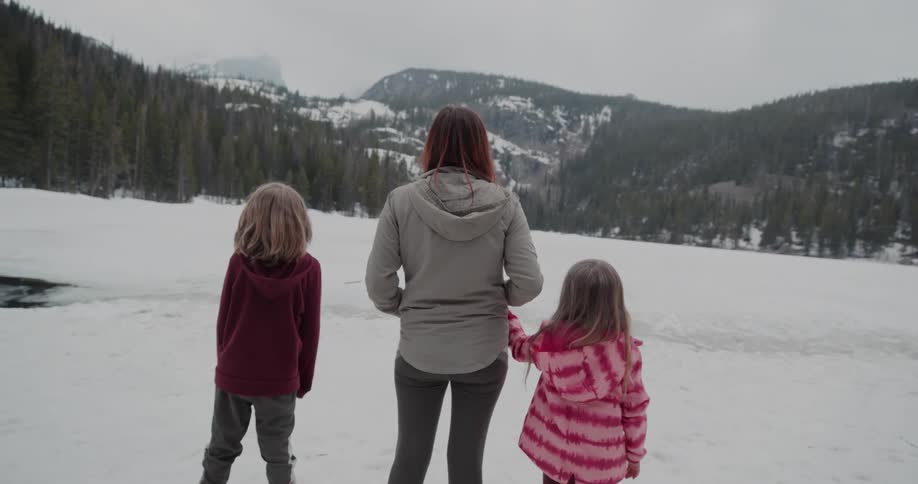 A woman with two children are standing outdoors in a snowy field while looking at the forest and mountains before them