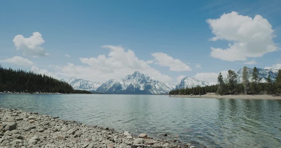 Water moves along the rock covered shore with forests on each side and snow covered mountains on the horizon