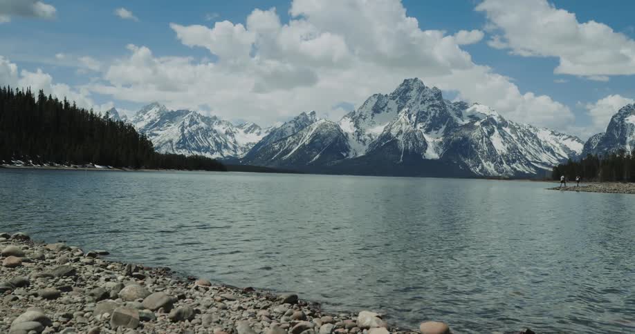 People are near a lake in front of snowy mountains on a cloudy day