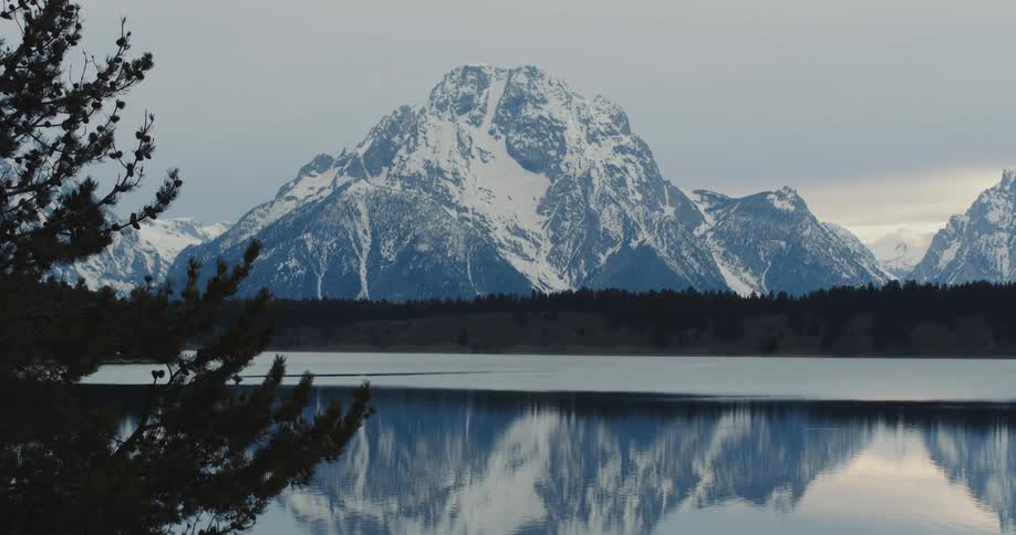 A lake surrounded by forest beneath a snowy mountain on a cloudy day
