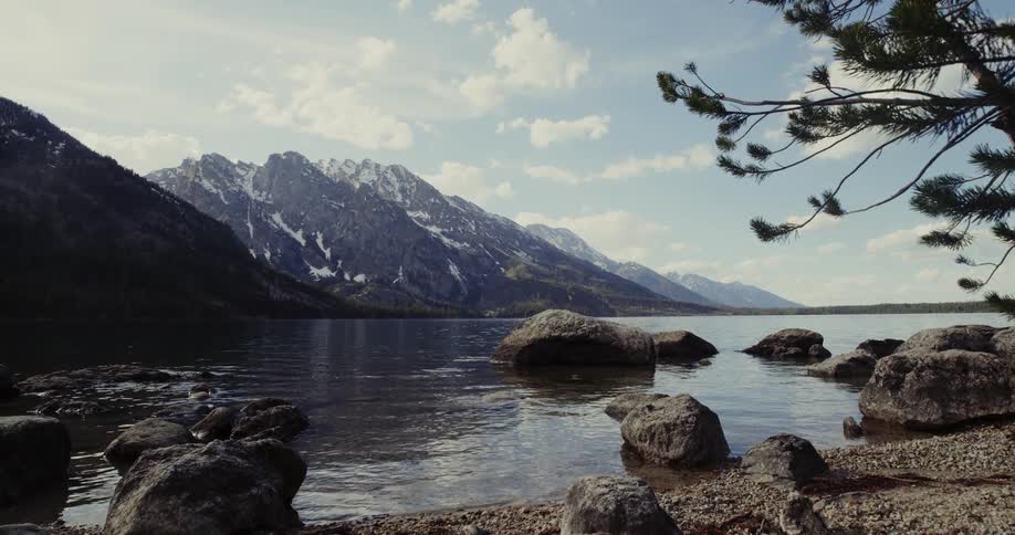 Water ripples around large stones near shore with snow dappled mountains in background