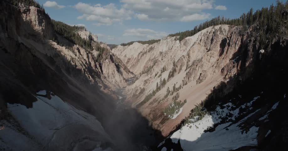 Conifer forest covers tops of mountain as river flows through snow coated mostly bare earth ravine