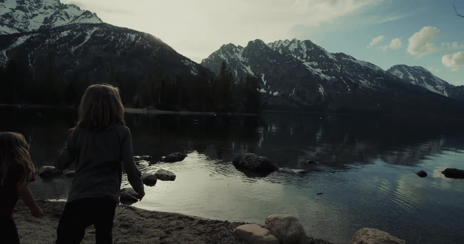 A child throwing a stone into a lake with a mountain range the sky and clouds in the background