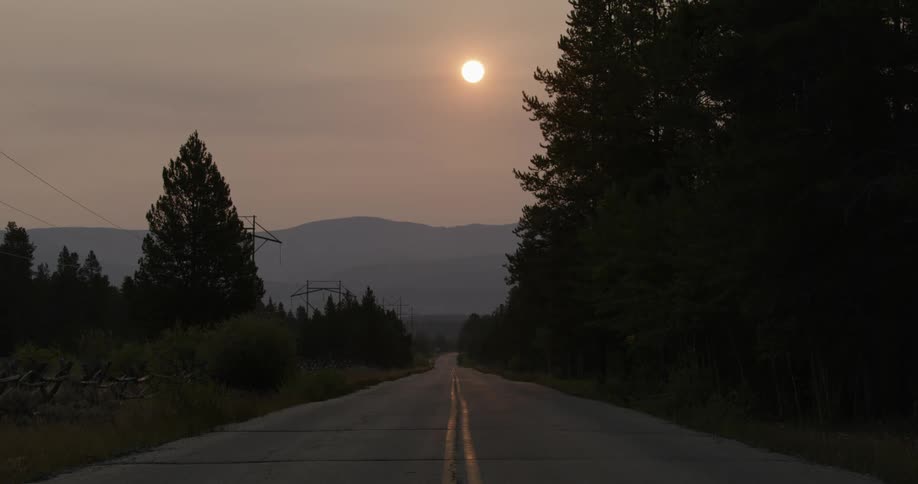 Sun shine through hazy from over misty mountains onto highway running through forest paralleling old wooden fence