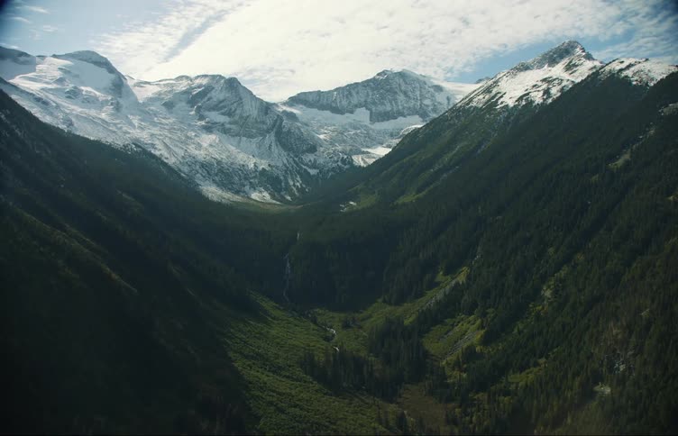 Aerial view of a valley with a river and a pine tree forest in front of the snow covered slopes of a mountain range