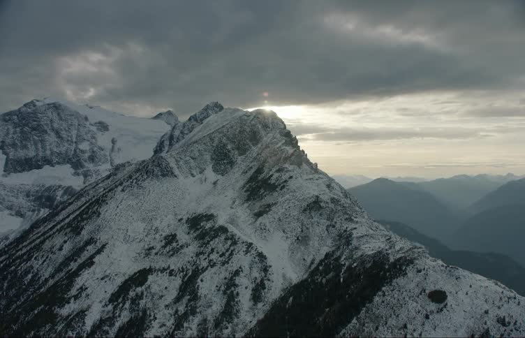 Sun shines through gray clouds onto snow covered mountain range and foggy valley