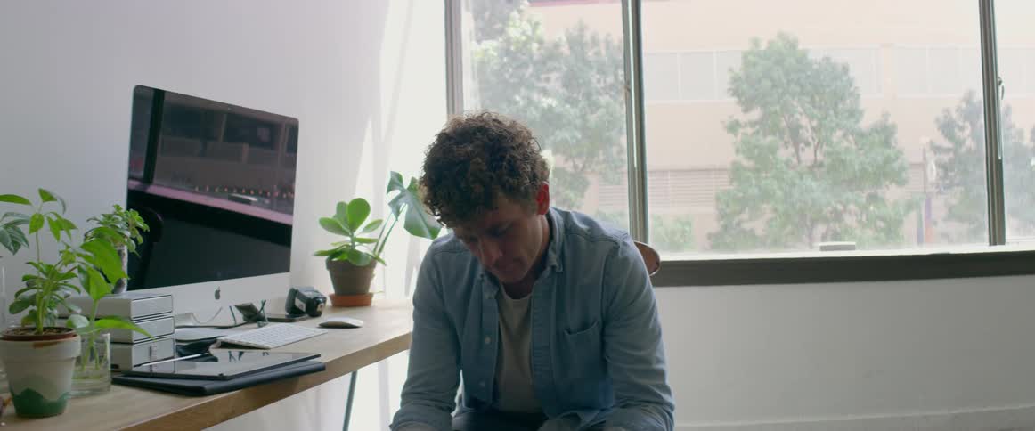A man is sitting in a room with a computer and large windows while looking down at something in his hands during the day