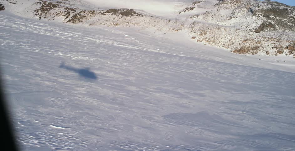 The ground covered in snow with the shadow of a helicopter with a cliff the sky and clouds in the background