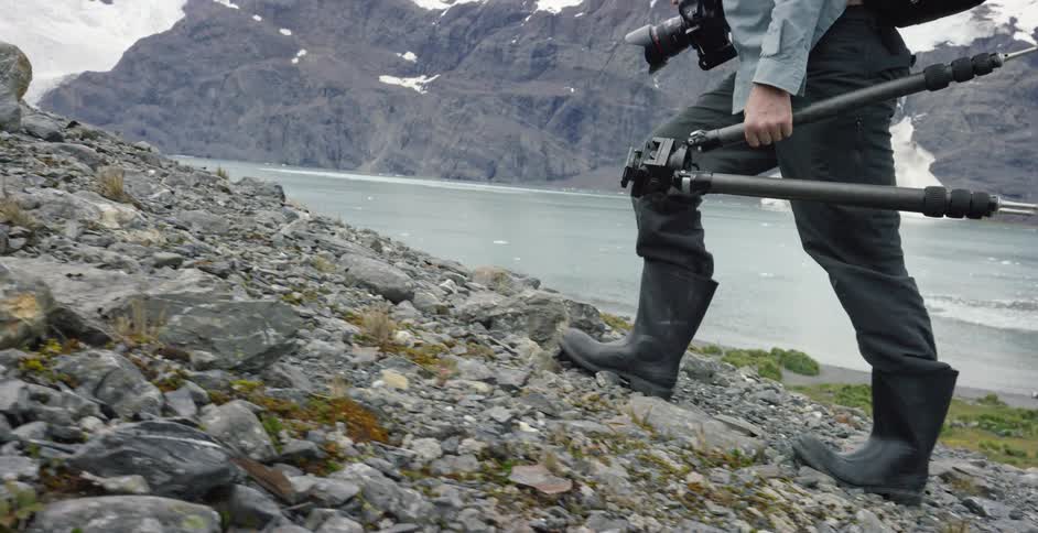 A man carries camera equipment up a rocky hill next to a river