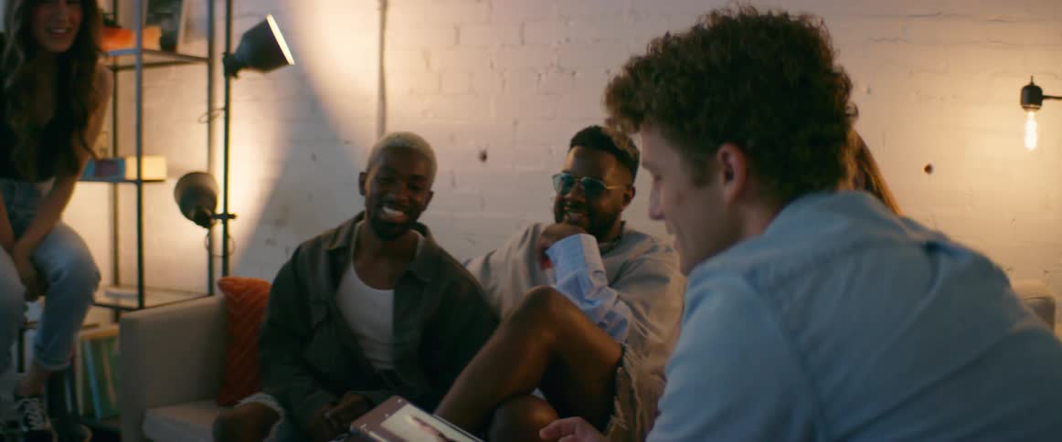 A group of friends sit in a house and sit and talk and laugh and look at pictures on a tablet