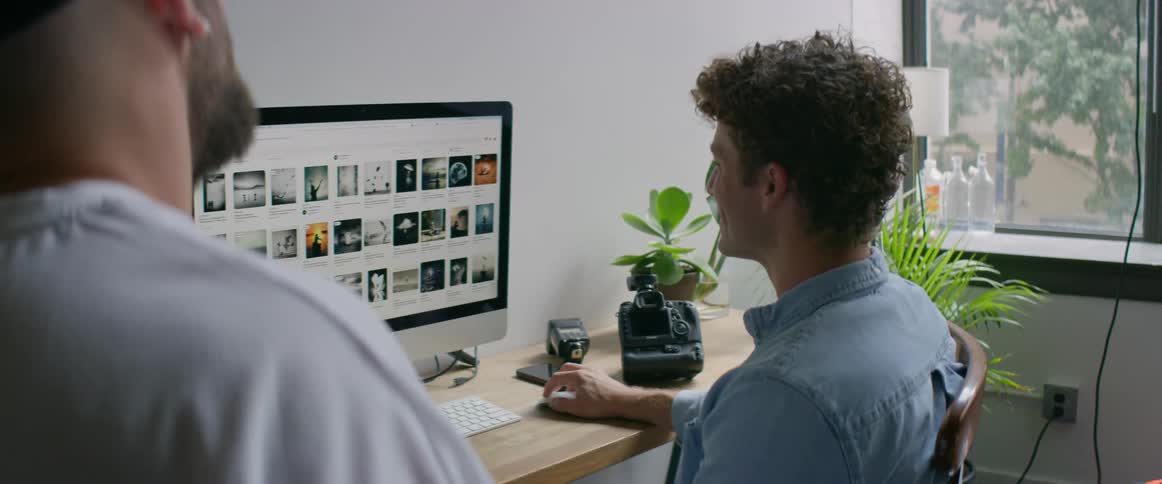 Two men in a room talking as once sits on a chair in front of a desk using a computer with a wall and window in the background