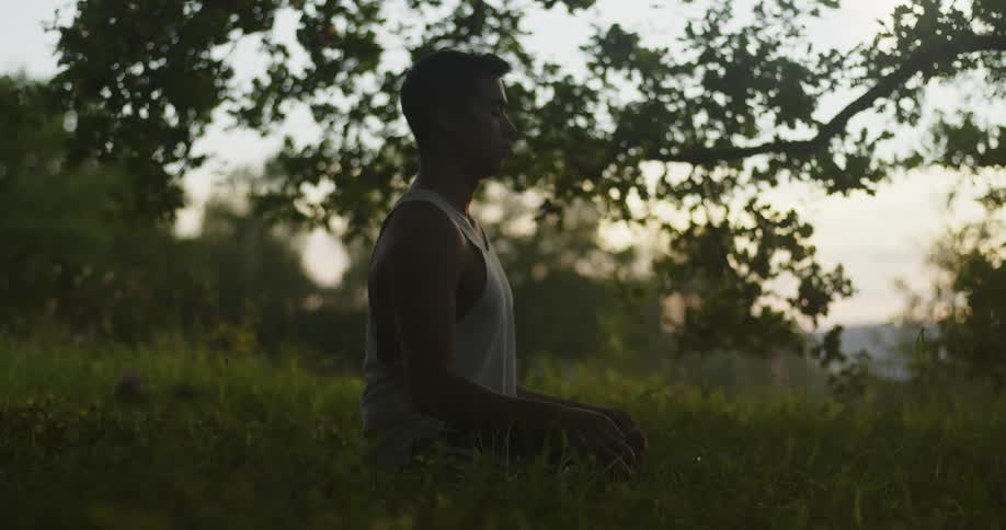 Young man on a white undershirt sitting on the grass in a yoga pose at evening