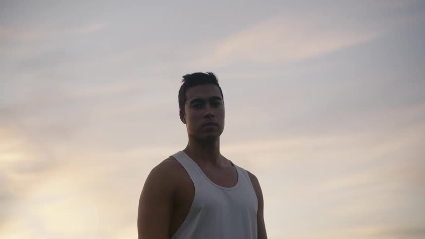 Man with white undershirt looking down at camera is silhouetted against the sky at dusk