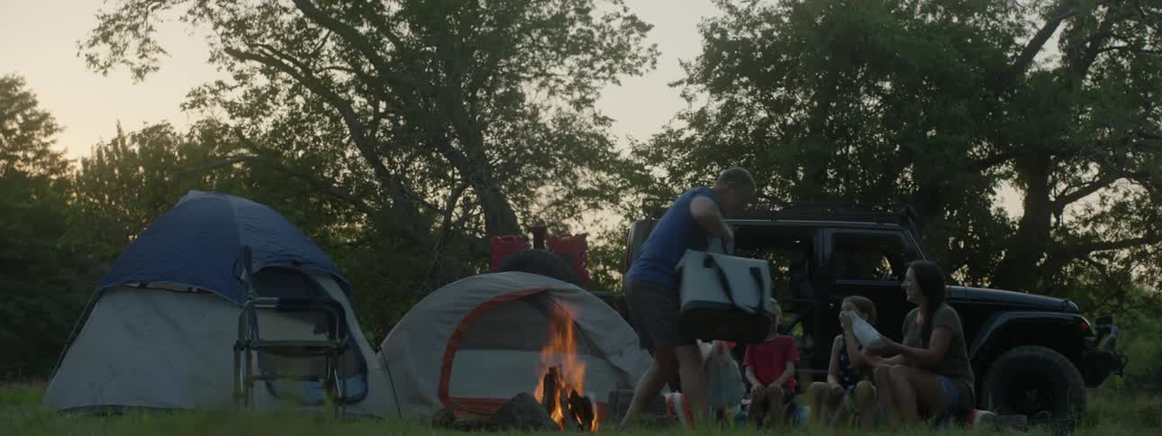 A man with bags in a campsite with a woman a boy and a girl with trees the sky and clouds in the background