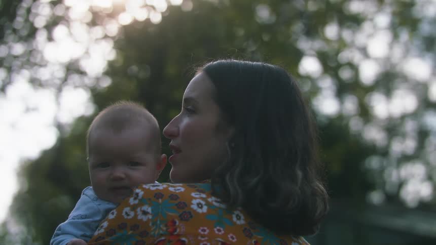 Mother with dark wavy hair orange blouse with flowers holds baby and the baby looks over mothers shoulder in the forest