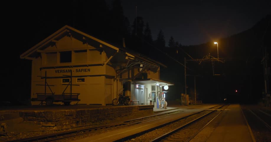 Train station emits light onto train tracks at night with conifer forest and mountain in background