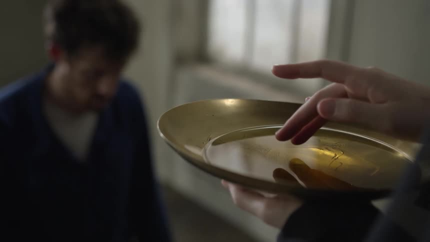 Person holding bowl dips hand into oil standing next to man with bowed head