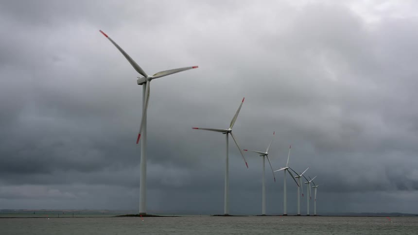 Spinning wind turbines along a river beneath a cloudy sky