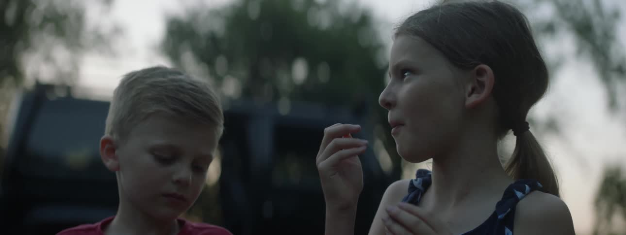 Father teases daughter and son as they stand eating and talking near parked vehicle
